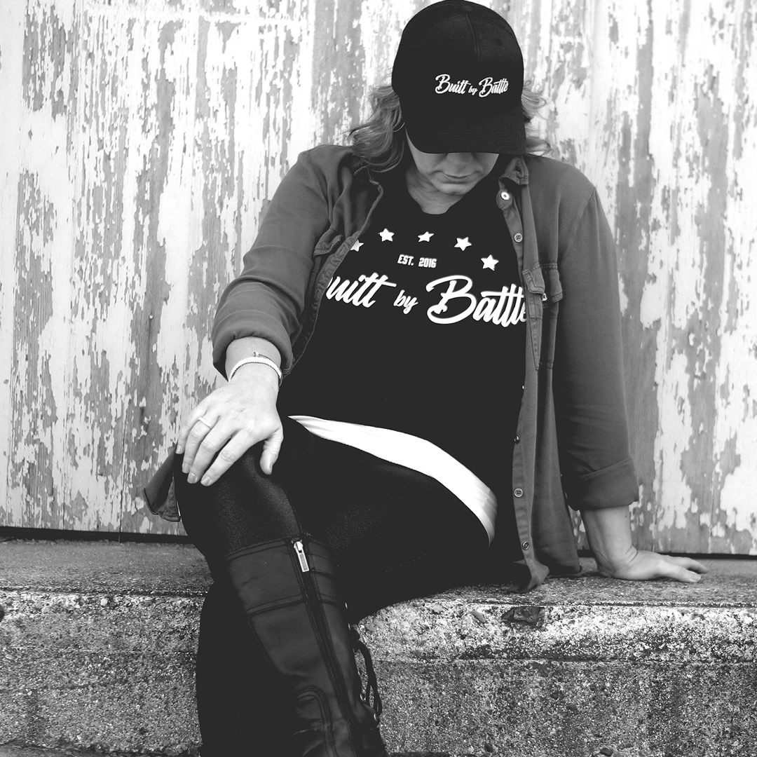 Woman in jeans and jacket sitting on concrete loading dock ledge with head down wearing Built By Battle t-shirt and hat